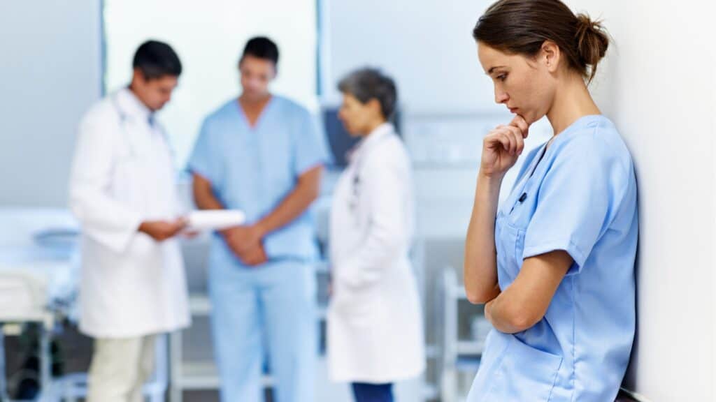 A nurse in blue scrubs stands apart from her colleagues, leaning against a wall with her hand on her chin, taking a quiet moment for herself while a team of doctors and a nurse confer in the background.